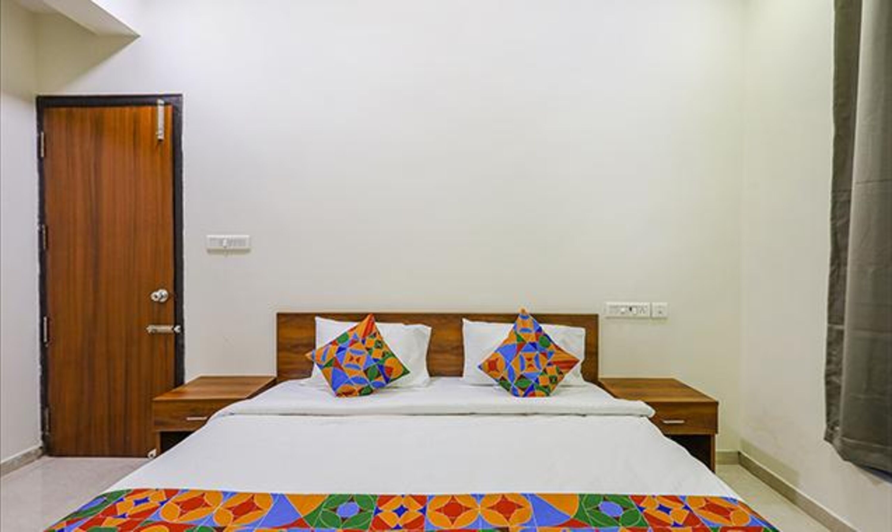 A bedroom with a wooden door, a bed with white bedding, two colorful pillows, and a patterned bedspread.