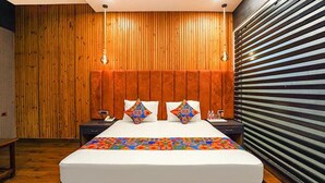 A modern hotel room with a wooden panel wall, a large bed with colorful bedding, and a striped blind on the window. - FabHotel Prime Tushar Elegant (Ghaziabad)