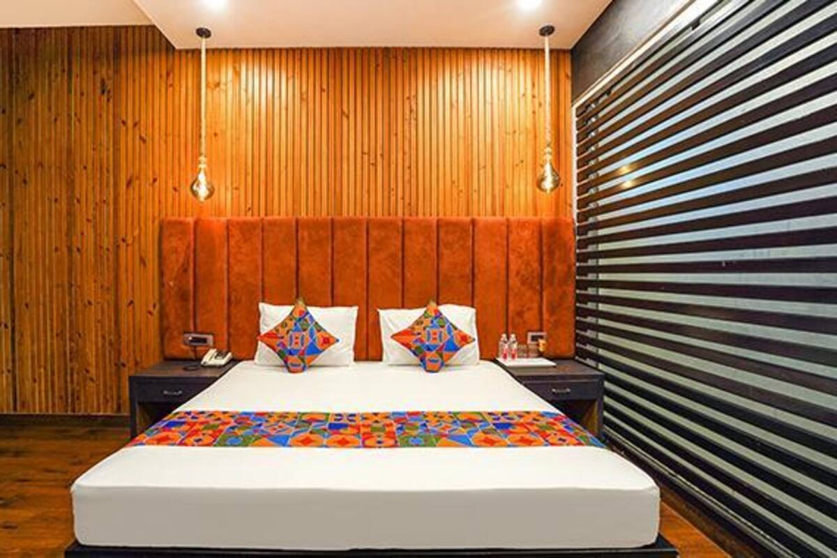 A modern hotel room with a wooden panel wall, a large bed with colorful bedding, and a striped blind on the window.