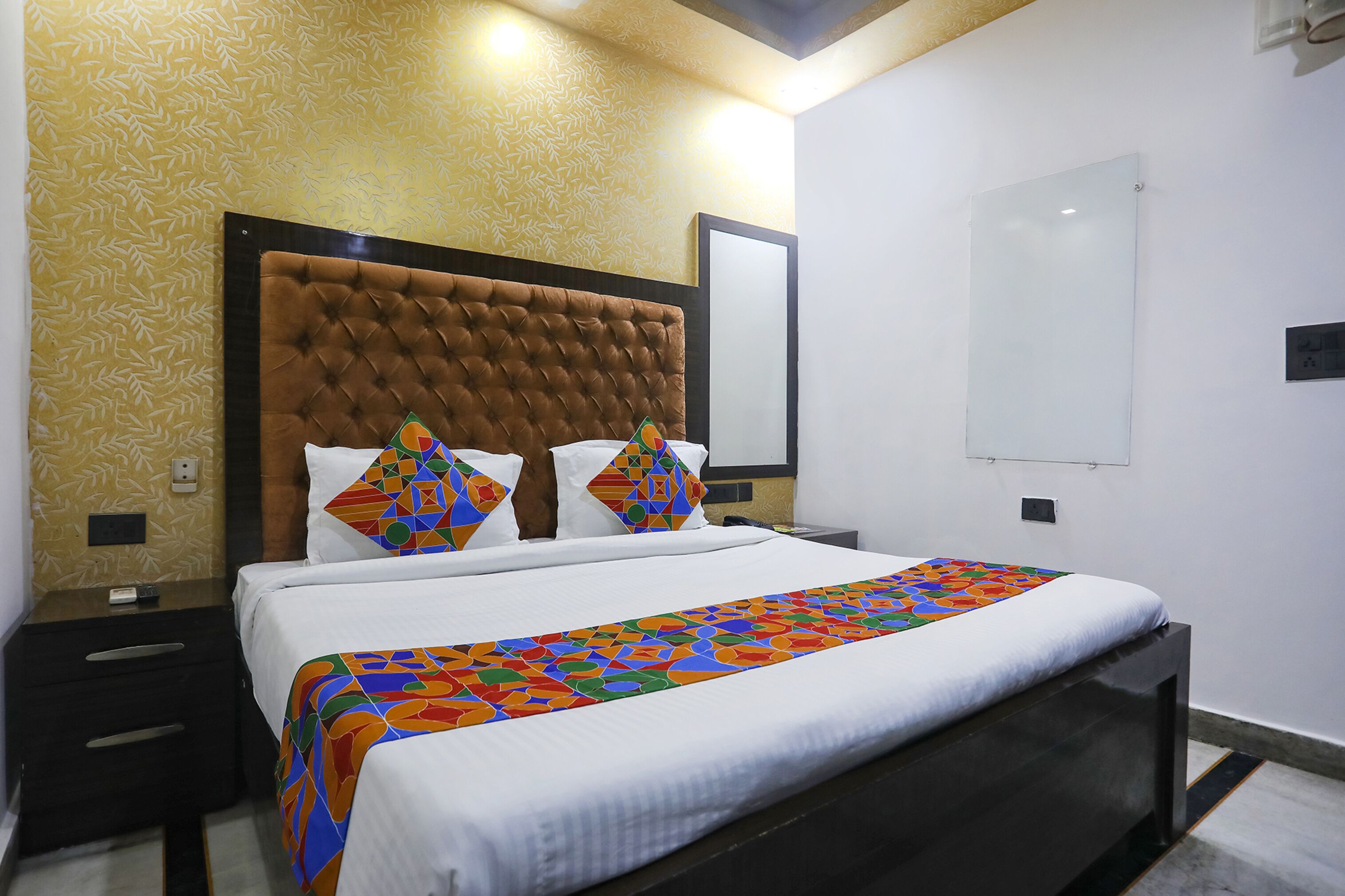 Une chambre d’hôtel avec une tête de lit capitonnée, un lit recouvert d’une couvre-lit aux motifs géométriques colorés et un mur sobre agrémenté d’un interrupteur.