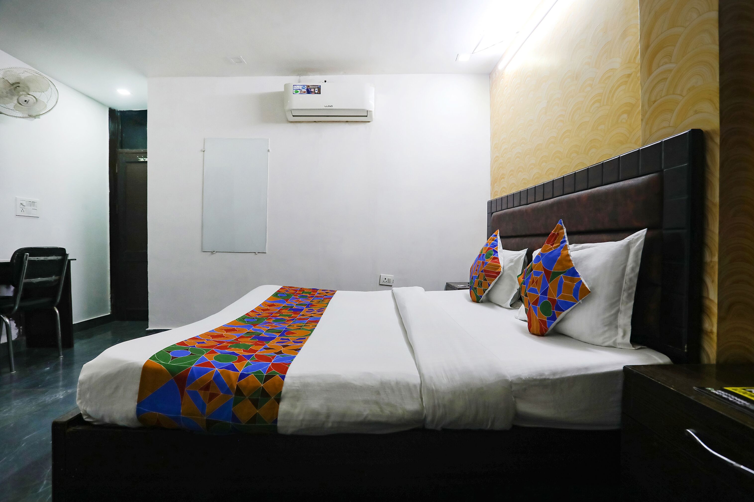 Una habitación de hotel con cama, mesita de noche, silla, aire acondicionado y ventilador de pared.