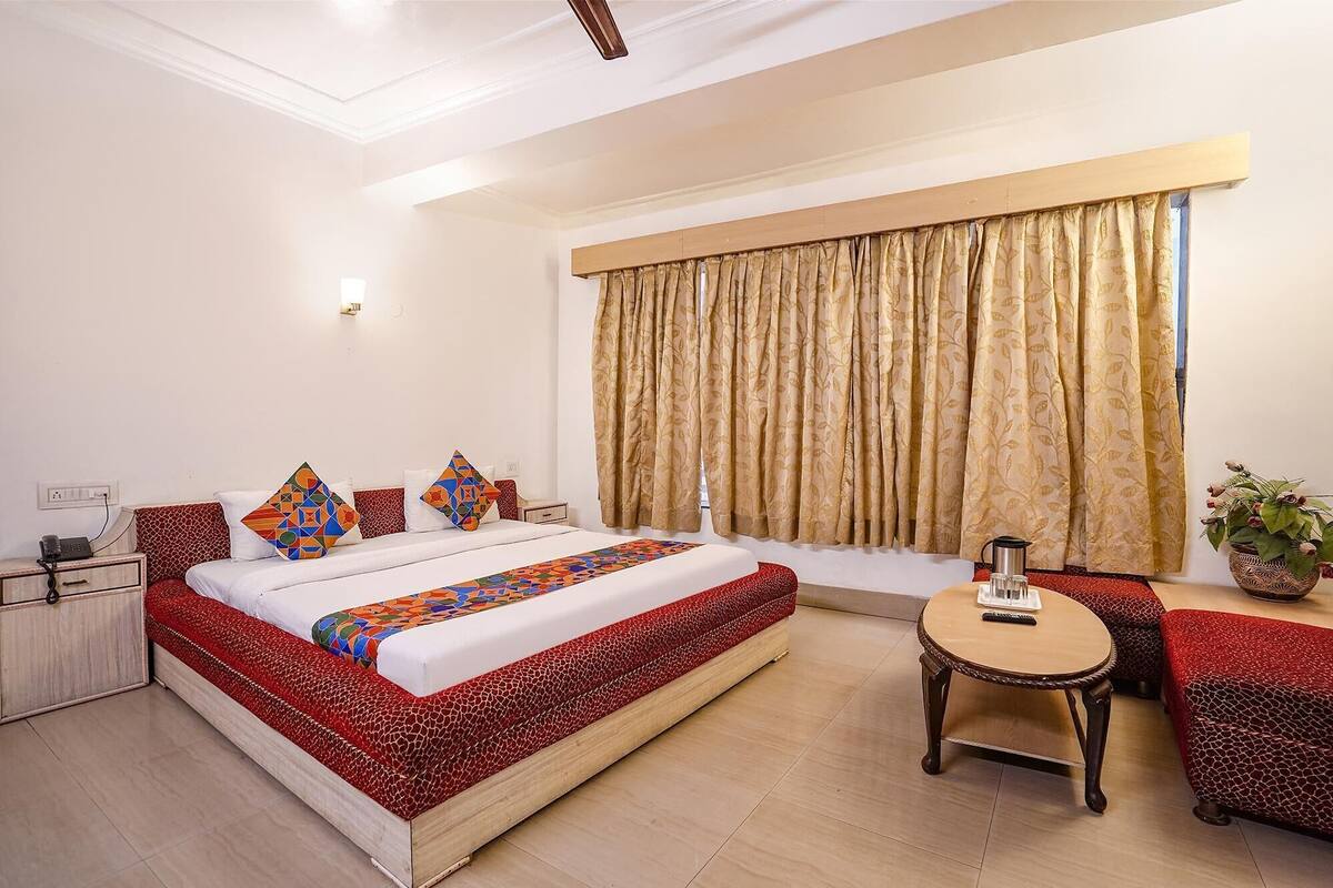 A bedroom with a large bed, a bedside table, a small table, a chair, a window with curtains, and a potted plant.