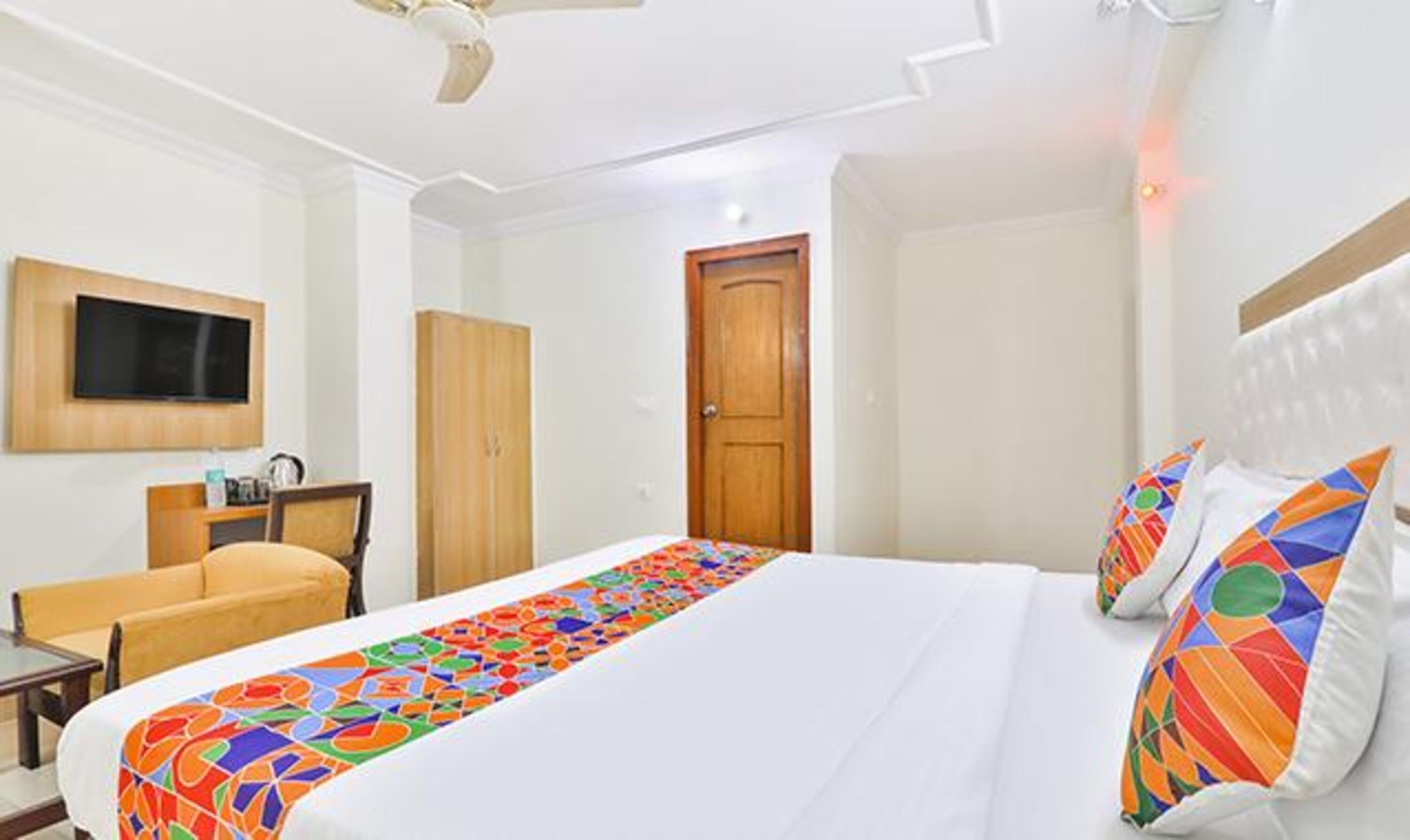 Habitación de hotel con cama, televisor montado en la pared, puerta de madera y ventilador de techo.