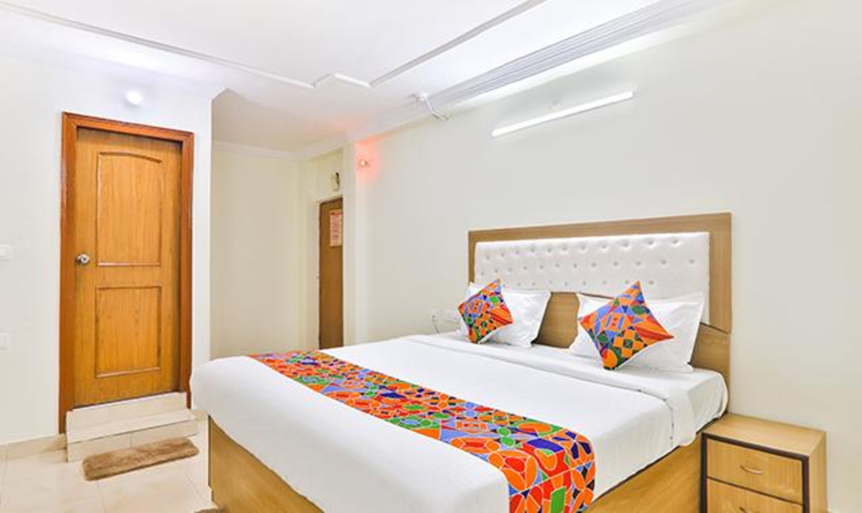 Habitació d'hotel amb una porta de fusta, un llit amb un edredó de colors, un panell de capçal blanc i una taula de nit amb un calaix.