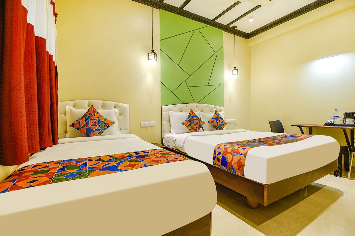 A hotel room with two beds, each with a colorful geometric-patterned bedspread and matching pillows. There is a green accent wall, a wooden headboard, and a small table with a water bottle and a coffee pot.