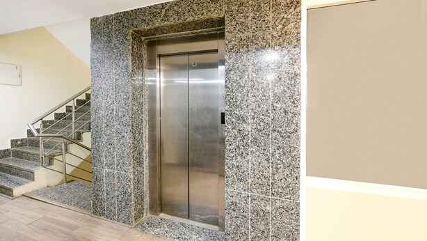 An elevator with a metallic finish, adjacent to a staircase with metal railings, and a tiled wall with a speckled pattern. - FabHotel Twamev (New Delhi)
