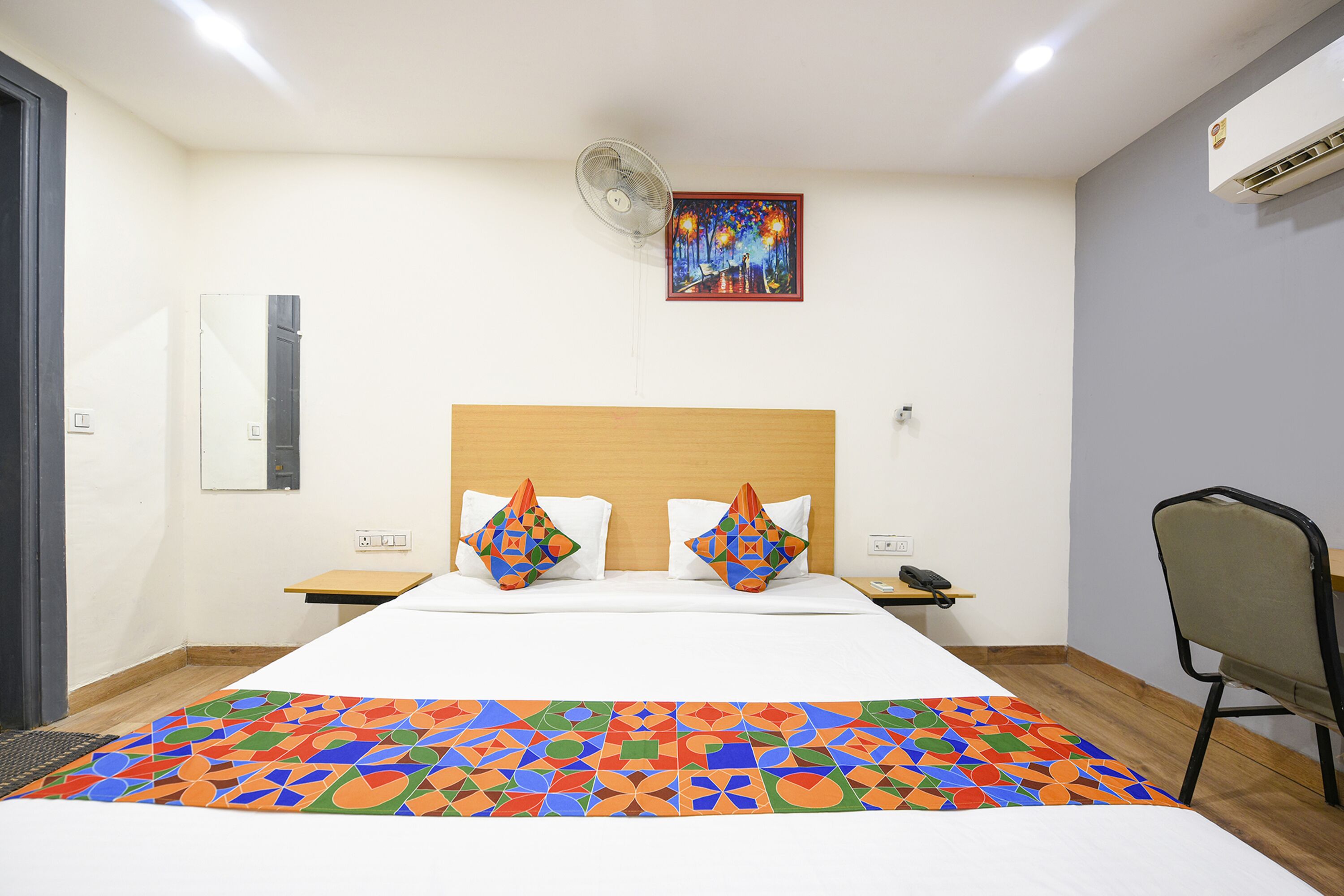 A hotel room with a bed, a nightstand, a chair, a wall-mounted air conditioner, and a ceiling fan.