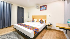 A hotel room with a bed, a bedside table, a nightstand, a desk, a chair, a window with curtains, a wall-mounted air conditioner, and a painting on the wall. - FabHotel Twamev (New Delhi)