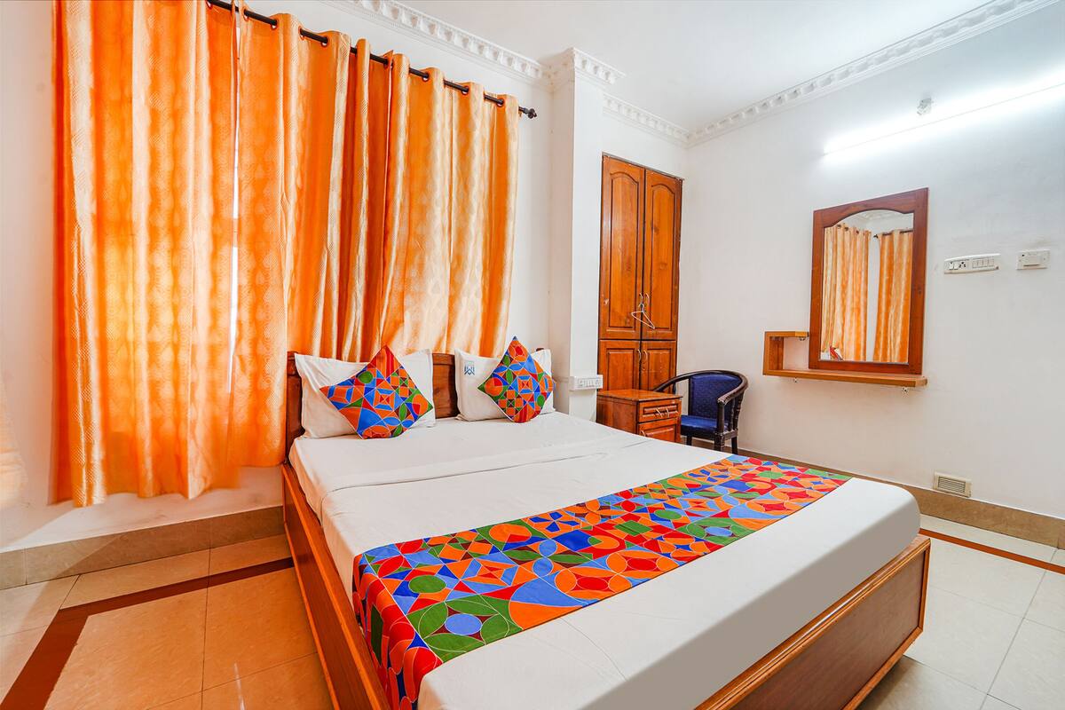 A hotel room with a bed, orange curtains, a wooden wardrobe, a chair, and a mirror on the wall.
