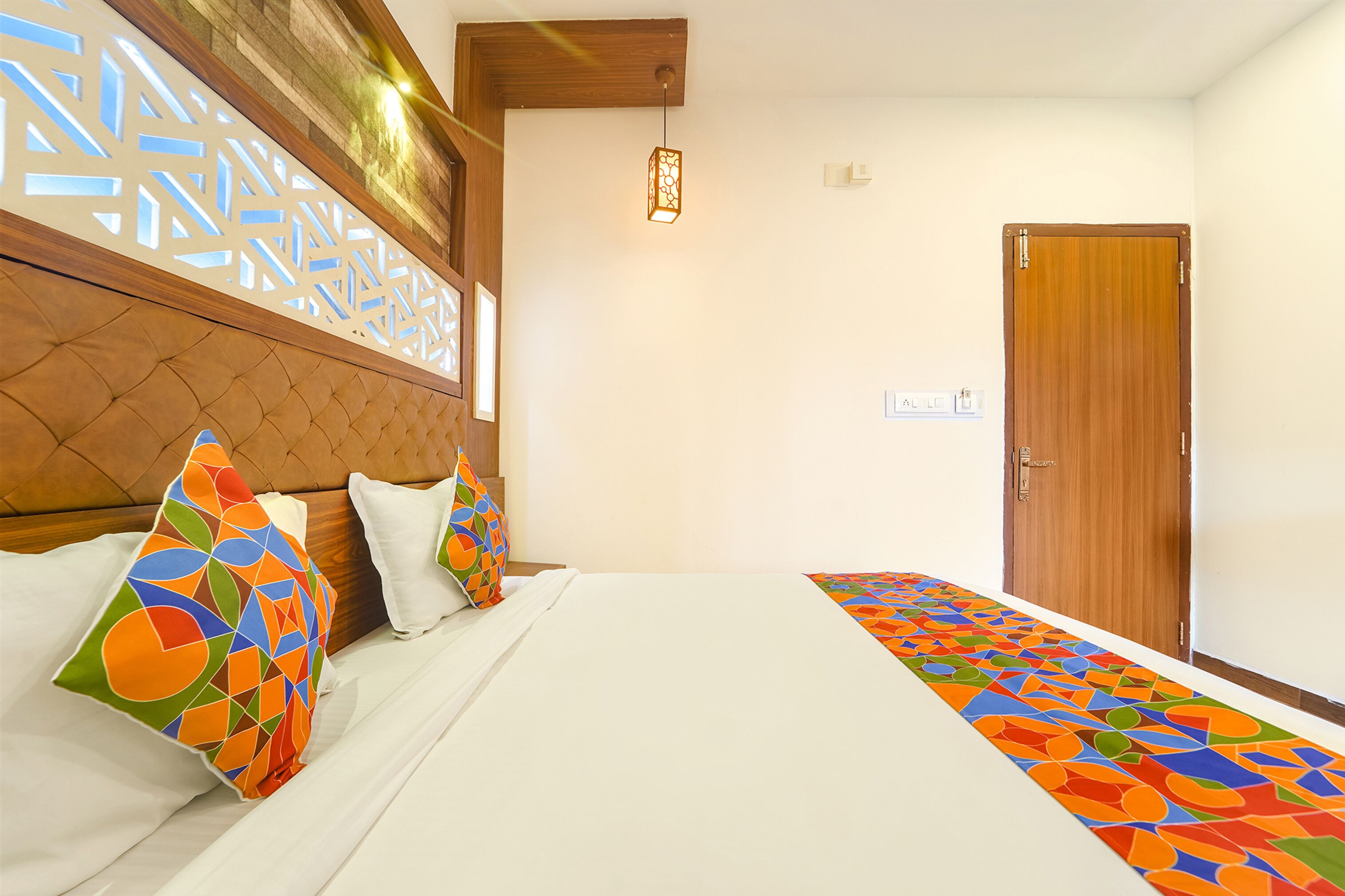 A hotel room with a bed, colorful pillows, a patterned headboard, a wooden door, and a hanging light fixture.