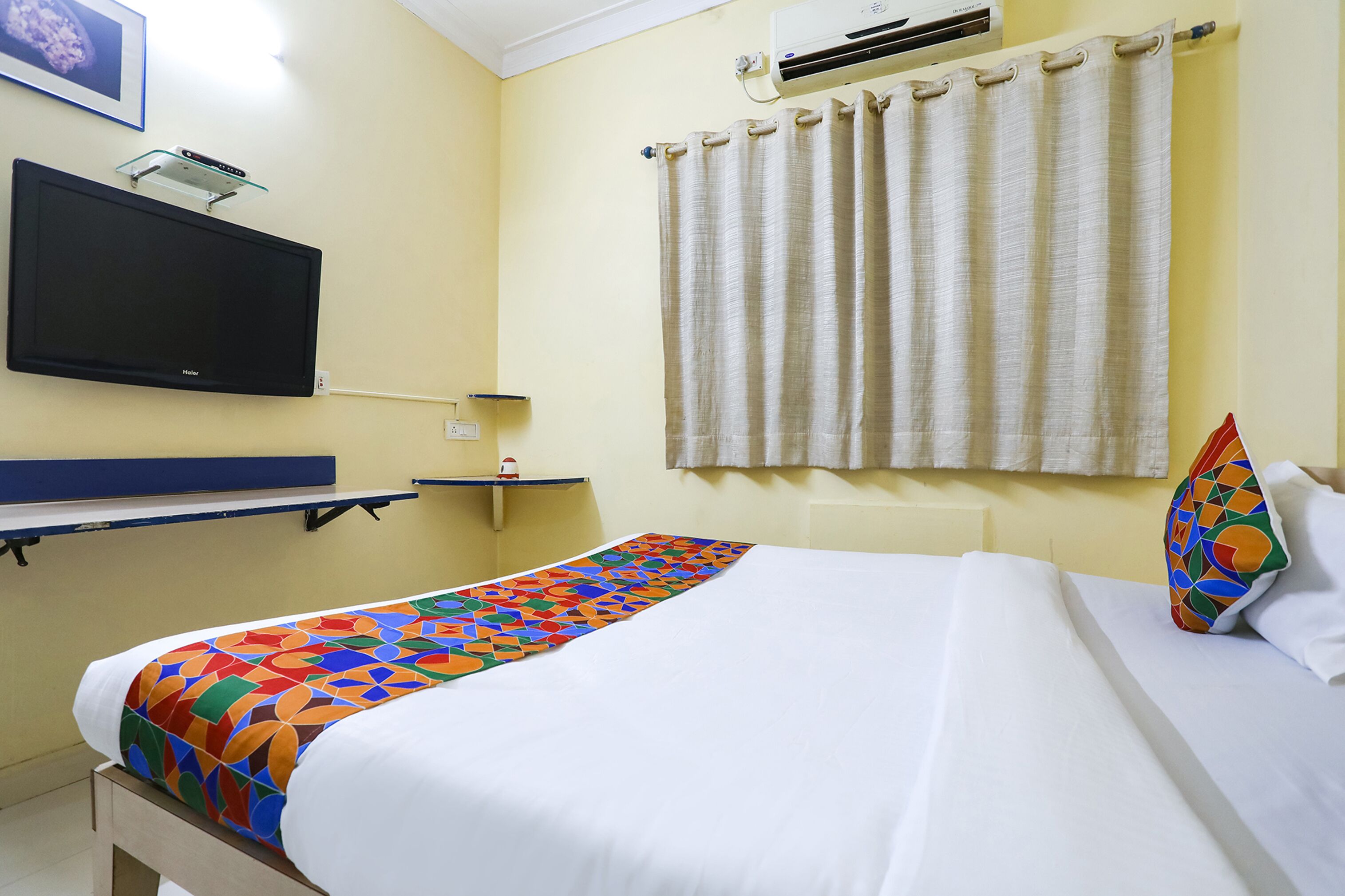 A hotel room with a bed, a flat-screen TV mounted on the wall, a window with curtains, and a small shelf with a lamp.