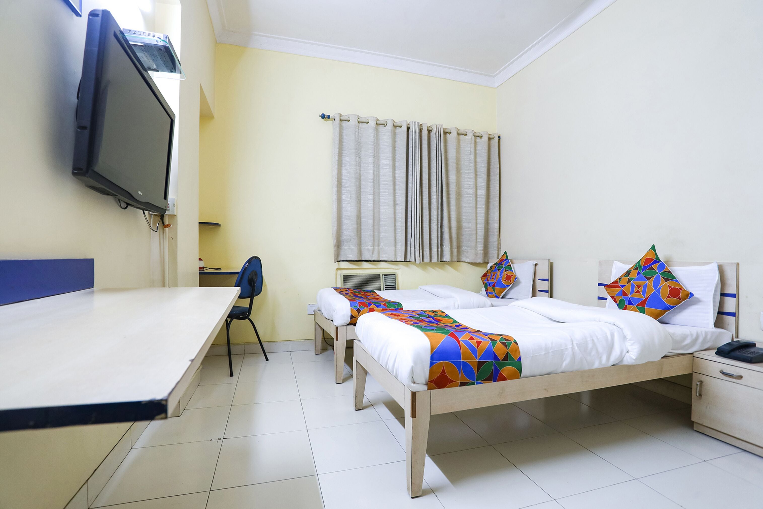 A hotel room with two single beds, a desk, a chair, a TV mounted on the wall, and a window with curtains.