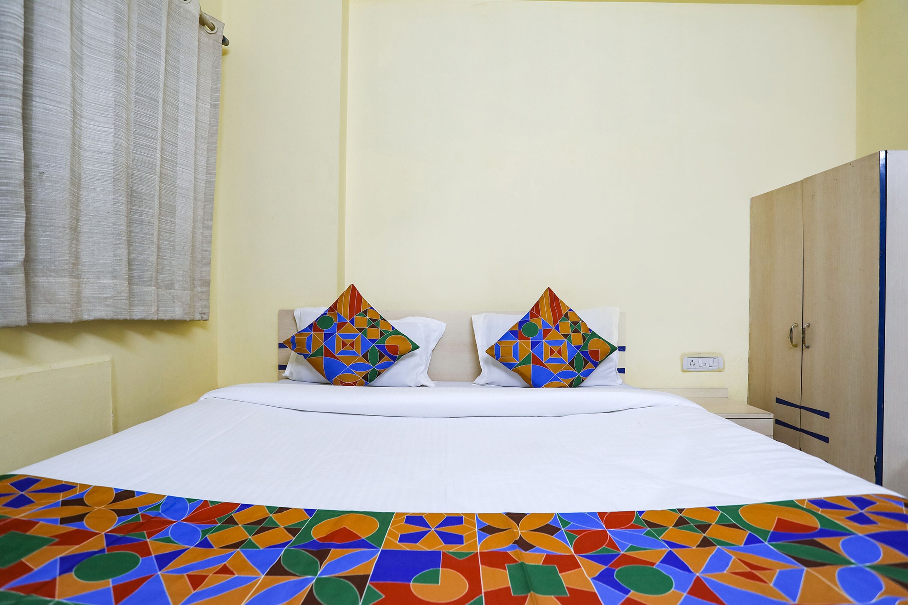 Een hotelkamer met een bed, twee kussens met geometrische patronen, een effen gordijn en een kledingkast.