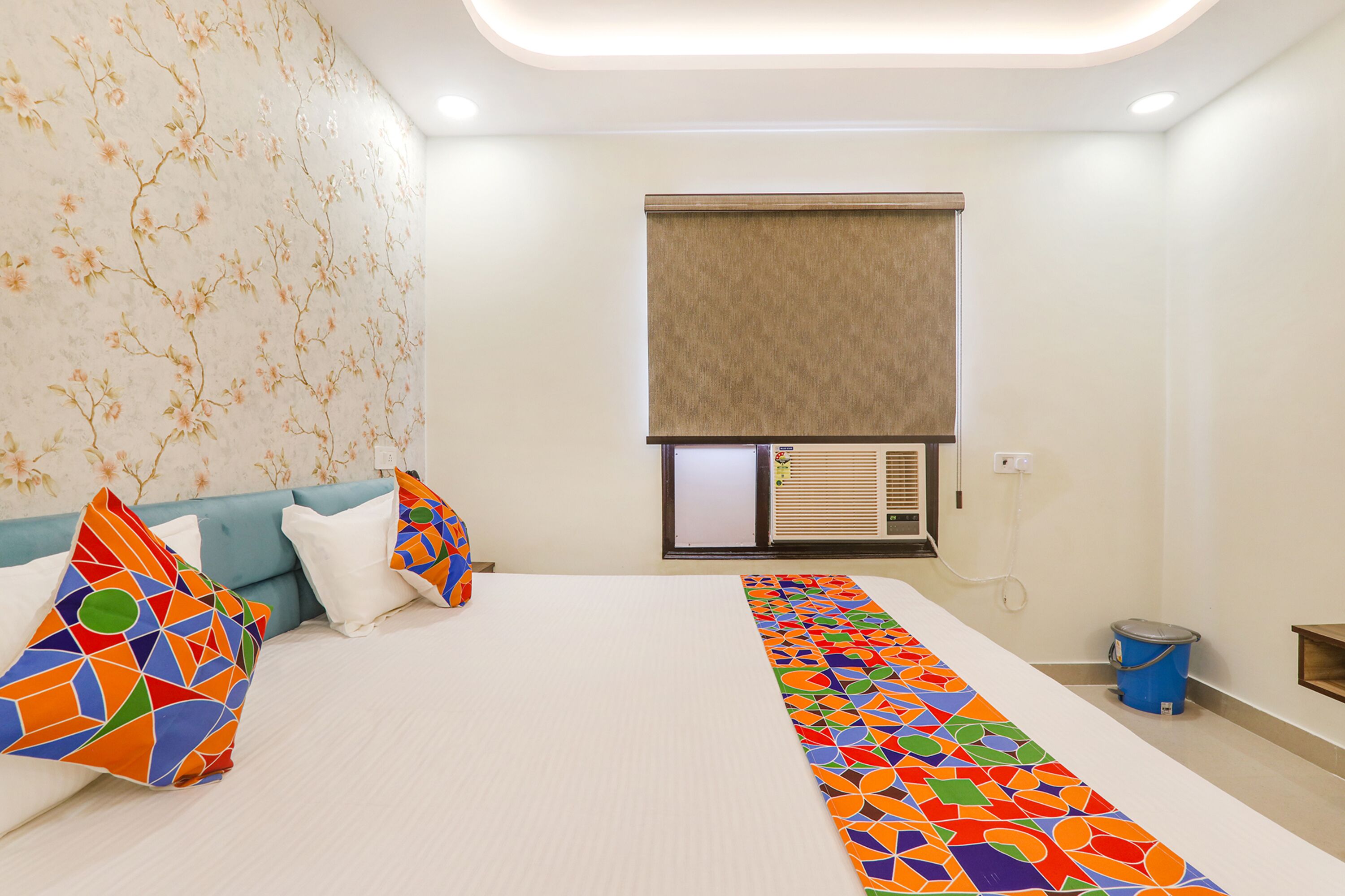 A hotel room with a bed, colorful pillows, a patterned runner, a window with blinds, an air conditioning unit, and a small trash bin.