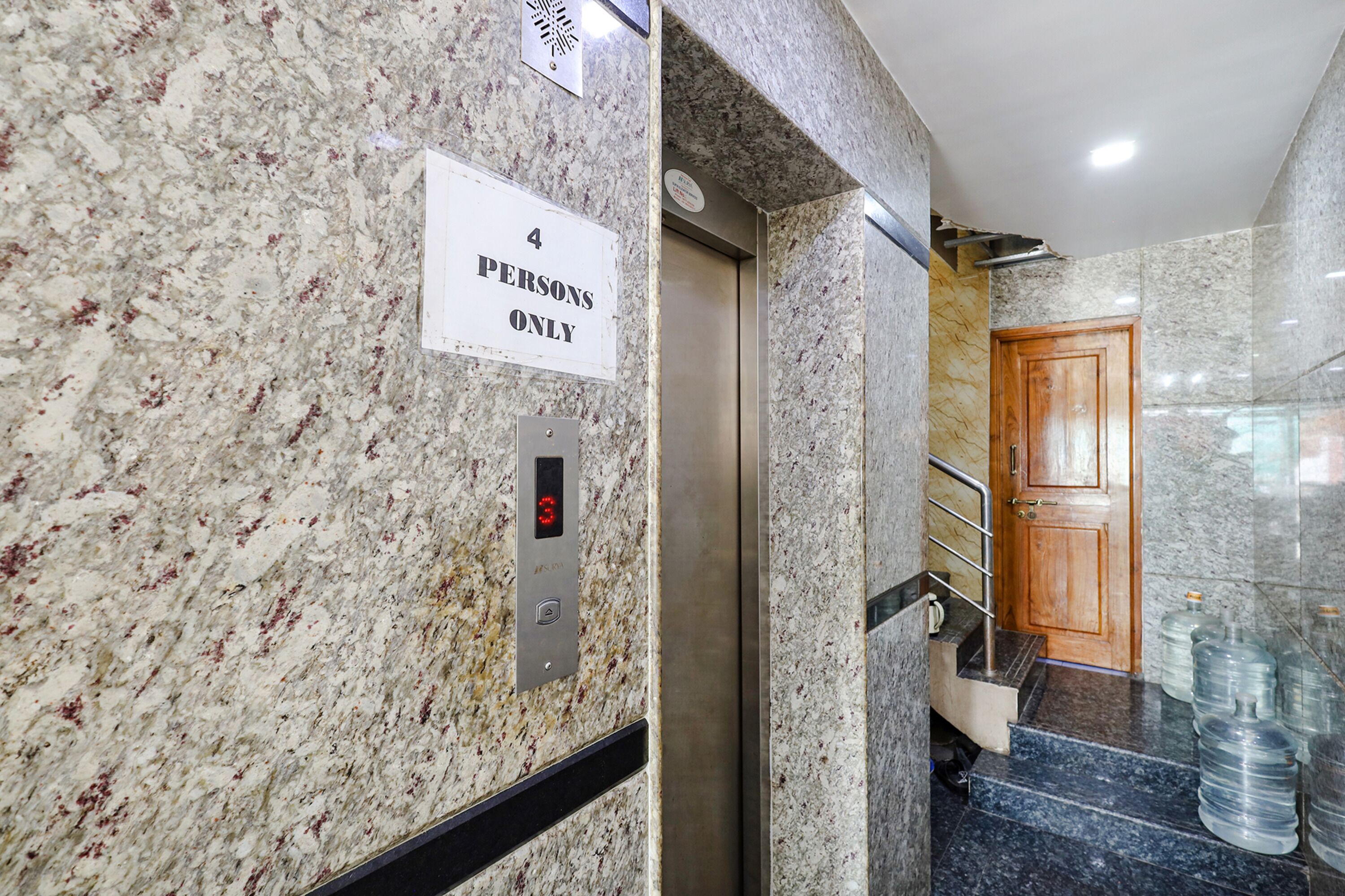 A marble wall with a '4 PERSONS ONLY' sign, an elevator, and a wooden door.