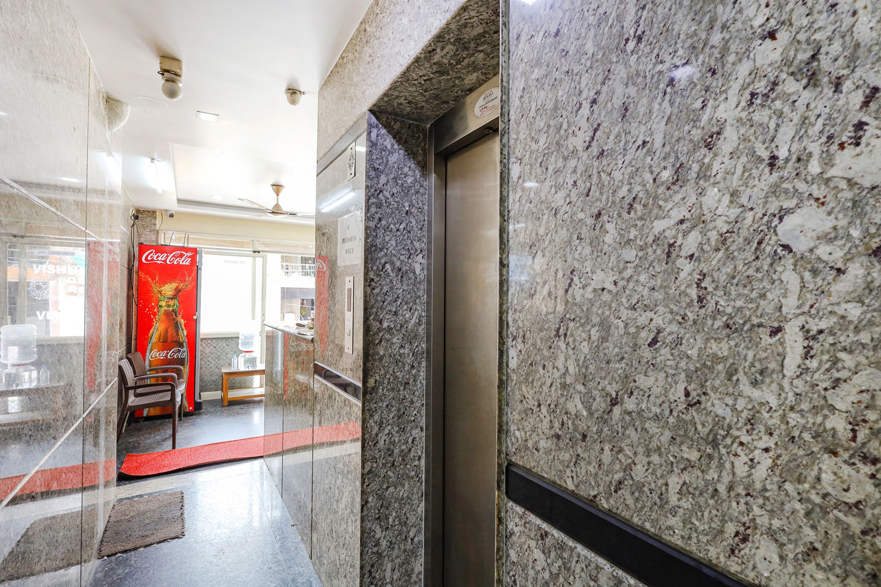 A modern lobby with a glass partition, a Coca-Cola advertisement, an elevator, and a reception area with a red carpet.