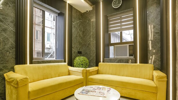 A yellow sofa and a small table in a room with marble walls and large windows. - Fabhotel National (Mumbai)