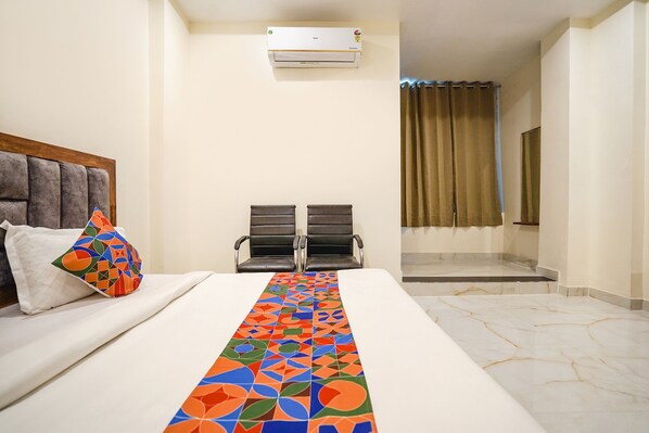 Une chambre d’hôtel avec un lit, un tapis coloré, deux chaises, un climatiseur et une vue sur une salle de bain équipée d’une douche et d’un rideau.