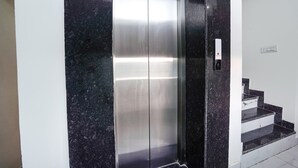 An elevator with a metallic finish, adjacent to a staircase with black steps and white walls. - Fabhotel Kyshna Inn (Mathura)