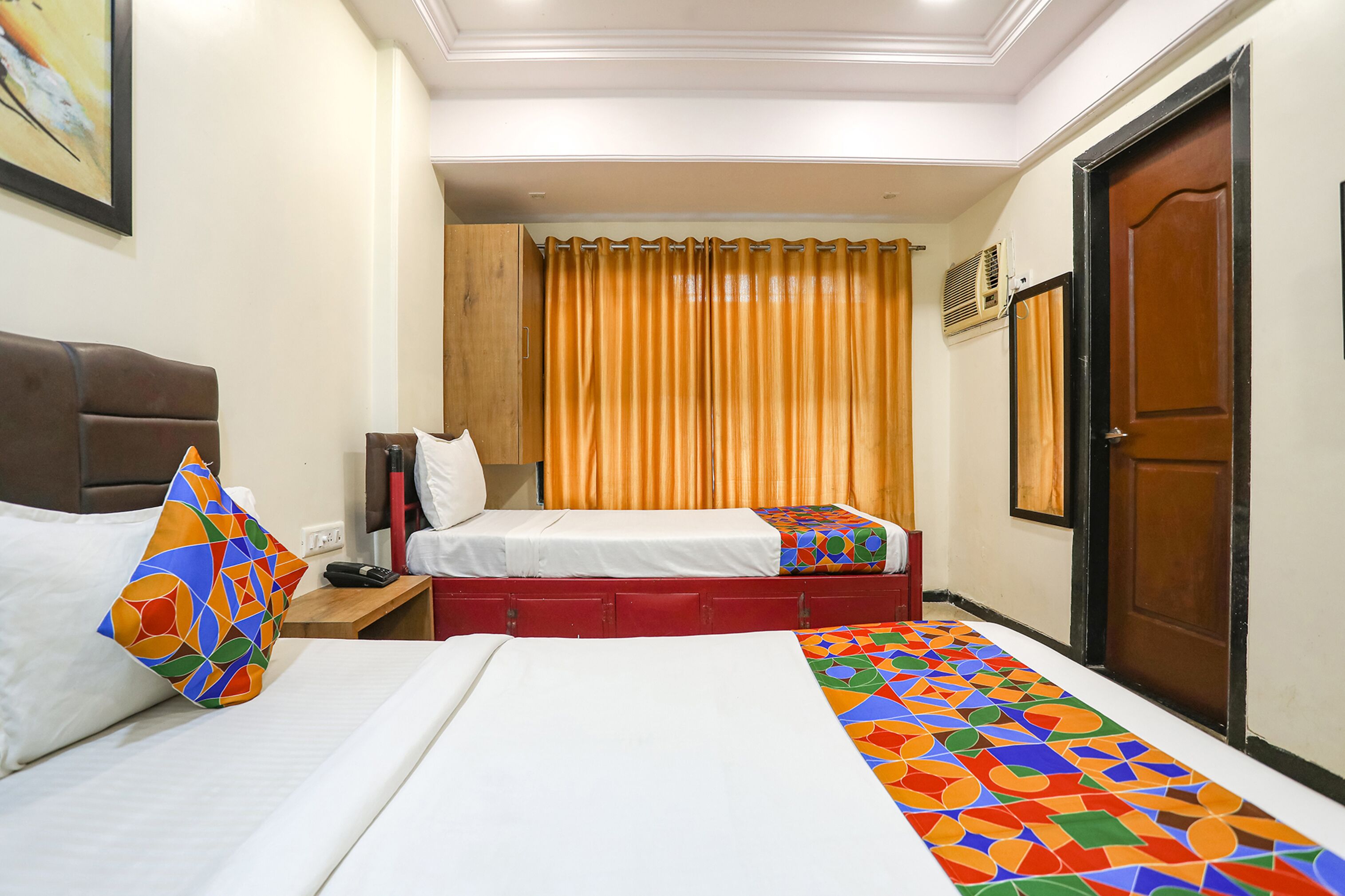 Habitación de hotel con dos camas, cabecera de madera, camino de cama colorido, mesita de noche y una puerta.
