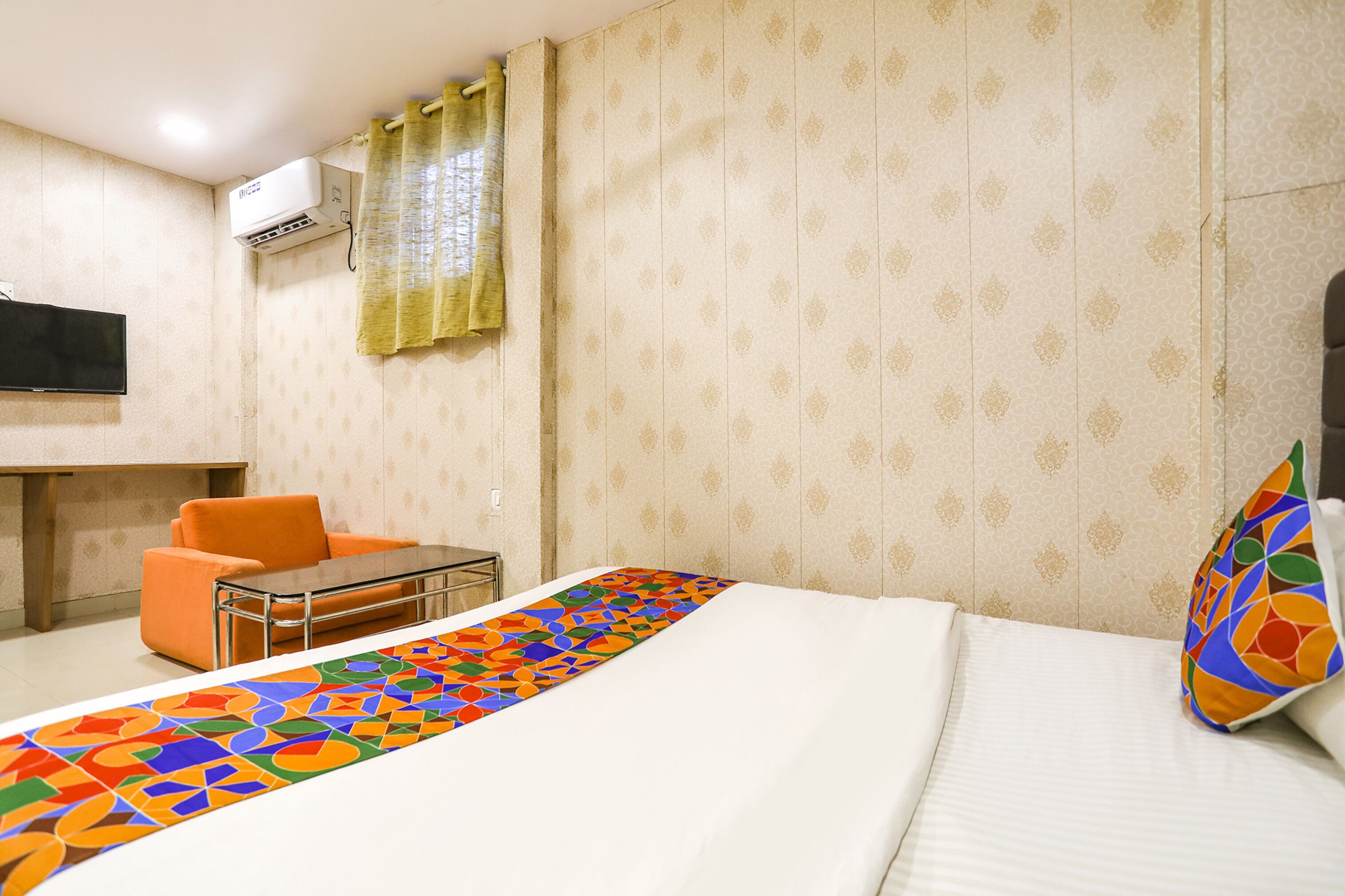 A hotel room with a bed, a small table, an orange chair, a TV, and an air conditioning unit.