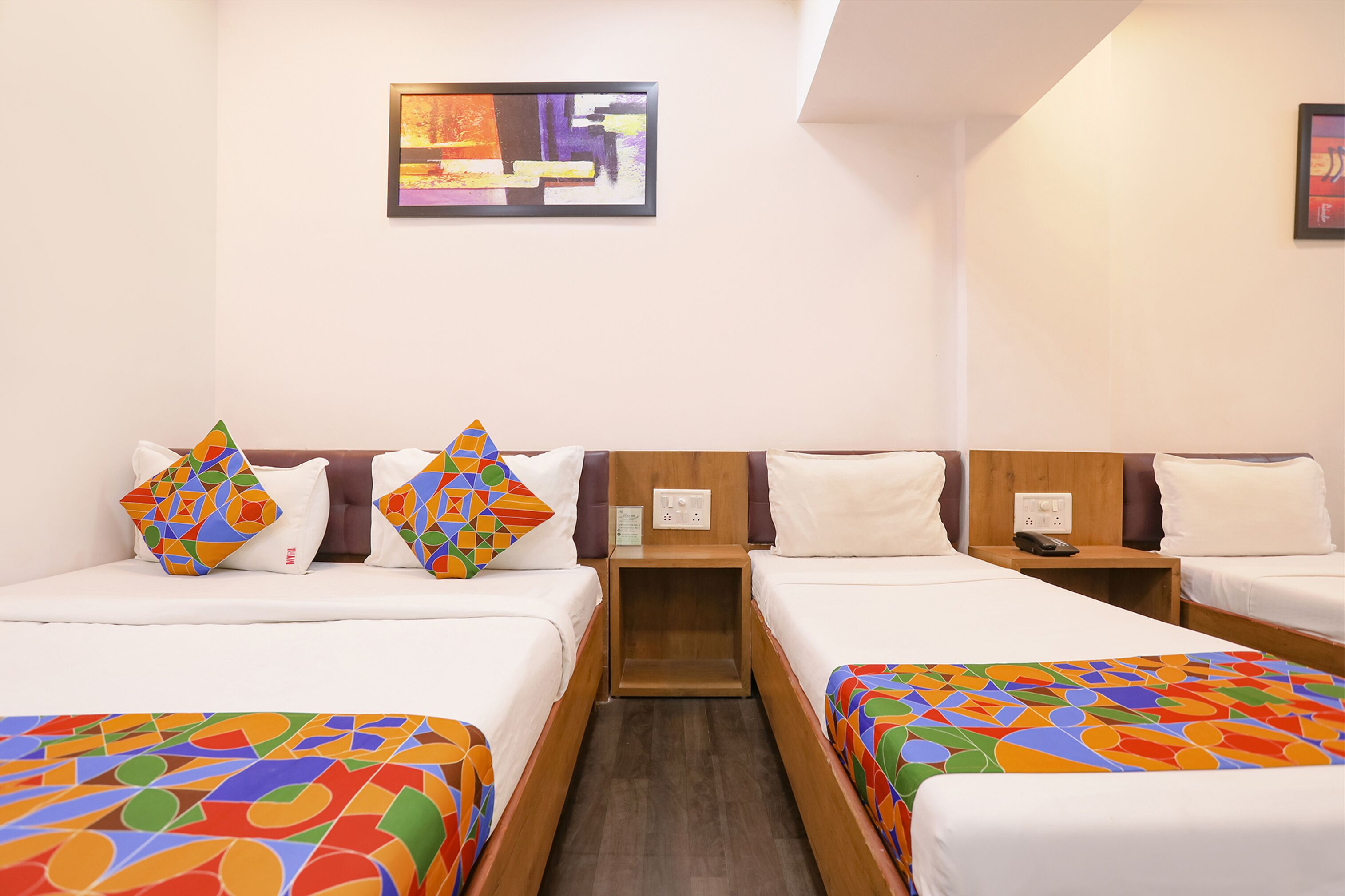 A hotel room with two single beds, each with a colorful geometric-patterned bedspread. There is a small wooden nightstand between the beds with a phone and a power outlet. The room has a simple, clean design with white walls and a single abstract painting on the wall.