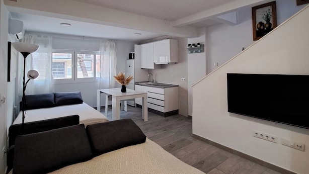 A modern hotel room with a bed, a desk, a kitchenette, and a TV.