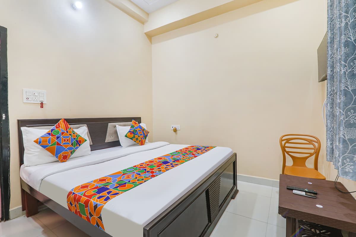A neatly made bed with a colorful geometric-patterned bedspread, a wooden headboard, and two pillows with geometric patterns. There is a small wooden table with a remote control on it, and a chair with a simple design.