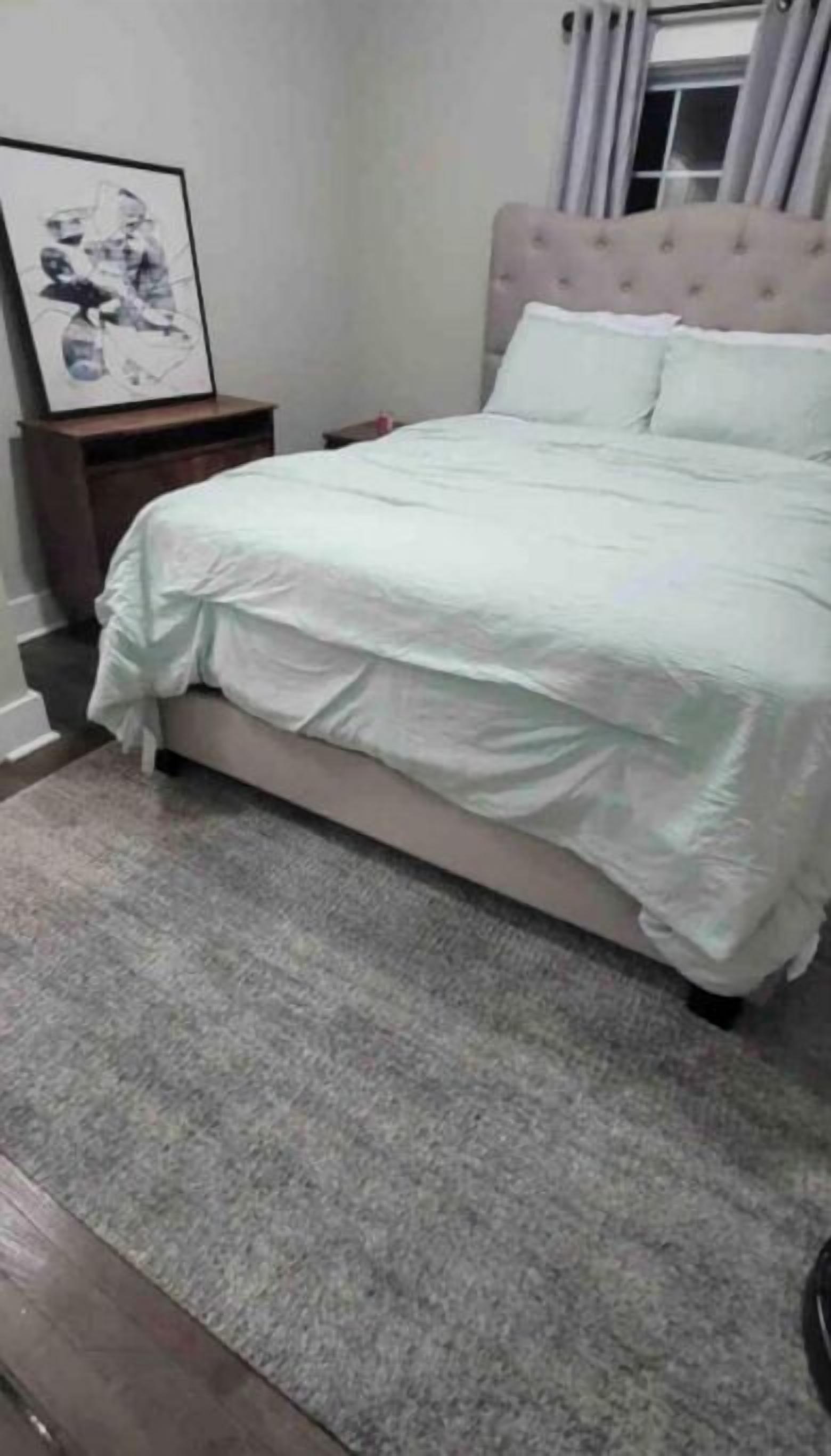 A bedroom with a bed, a nightstand, a framed picture, and a window with curtains.