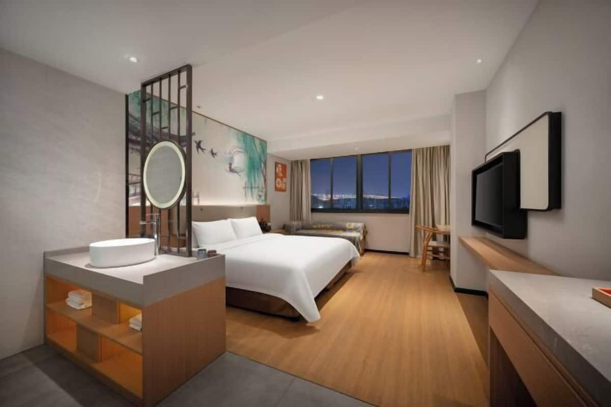A modern hotel room with a bed, a vanity with a mirror, a television, and a view of the city at night.