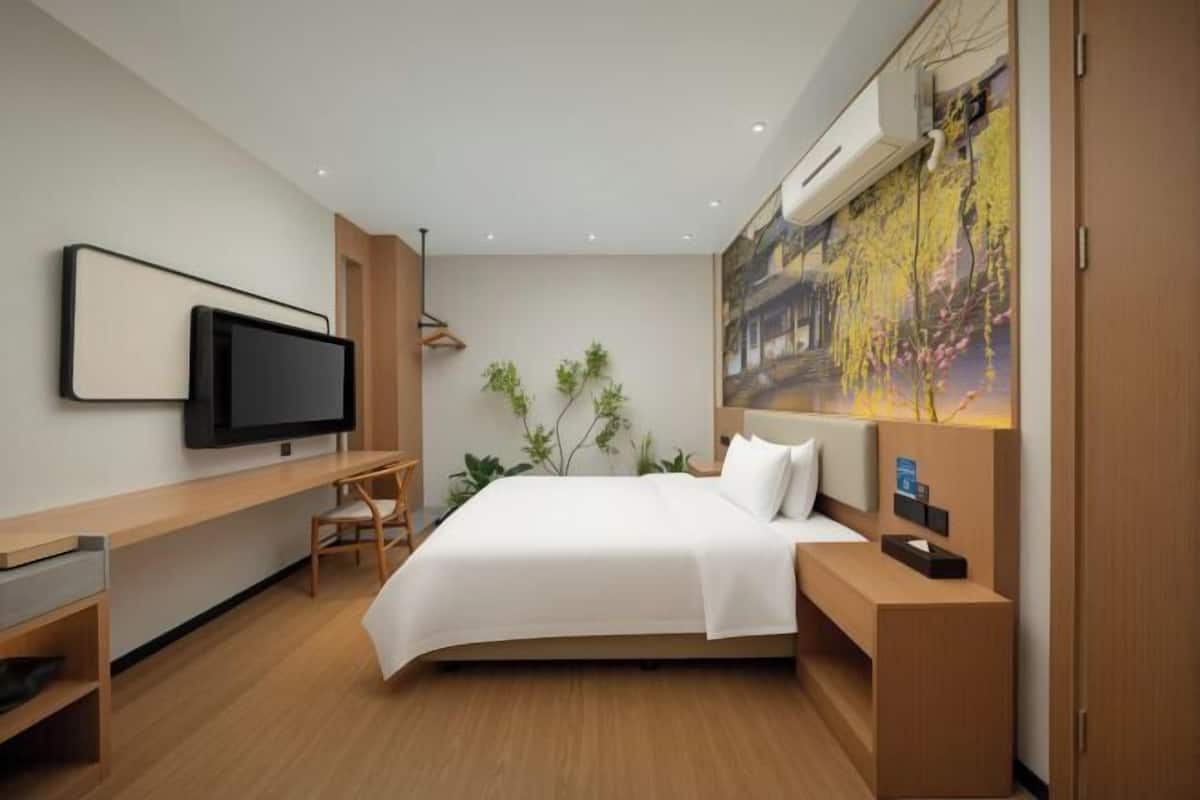 A modern hotel room with a bed, a desk, a television, and a large painting on the wall.
