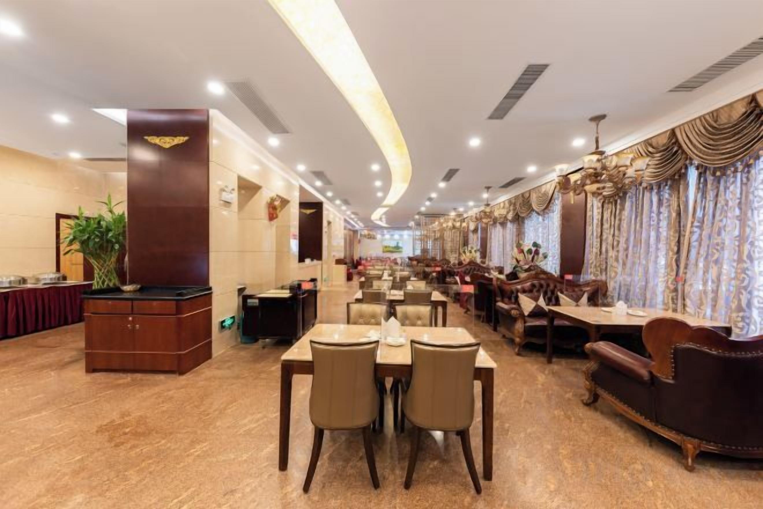 A spacious hotel dining area with multiple tables and chairs, a reception desk, and a chandelier.