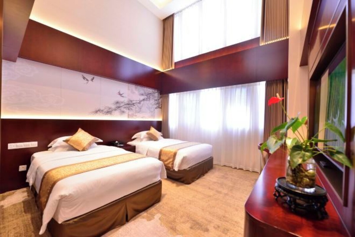 Une chambre d’hôtel avec deux lits, une grande fenêtre, une œuvre d’art accrochée au mur et une plante posée sur une table.