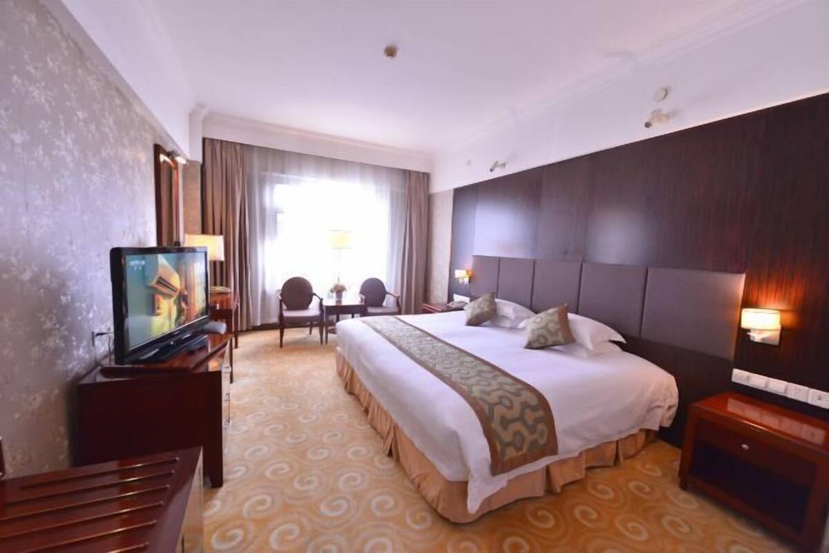 Hotelkamer met een groot bed, een televisie op een standaard, een bureau met een stoel en een raam met gordijnen.