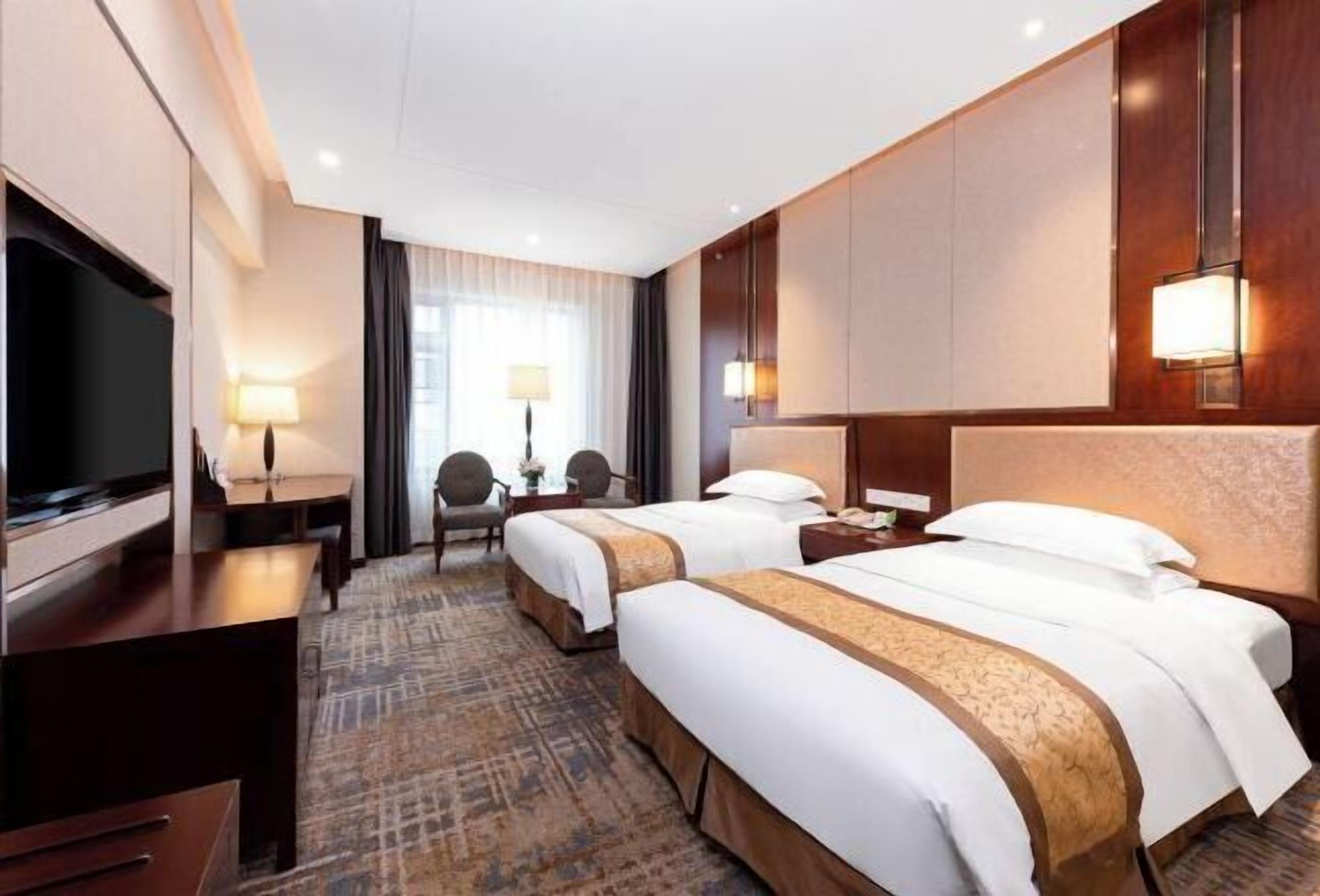 A hotel room with two beds, a TV, a desk with a lamp, and a view of the outdoors.