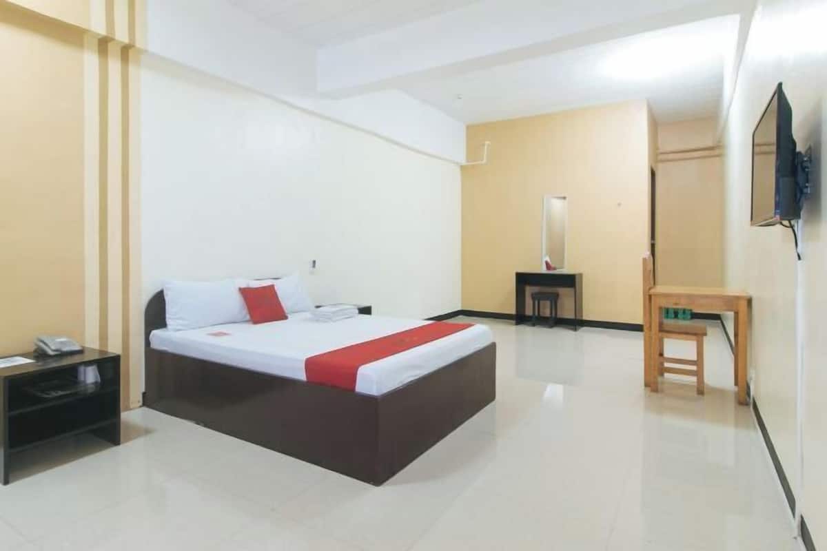 Ein Hotelzimmer mit Bett, Nachttisch, einem kleinen Holzhocker, einem an der Wand montierten Fernseher und einem Spiegel.