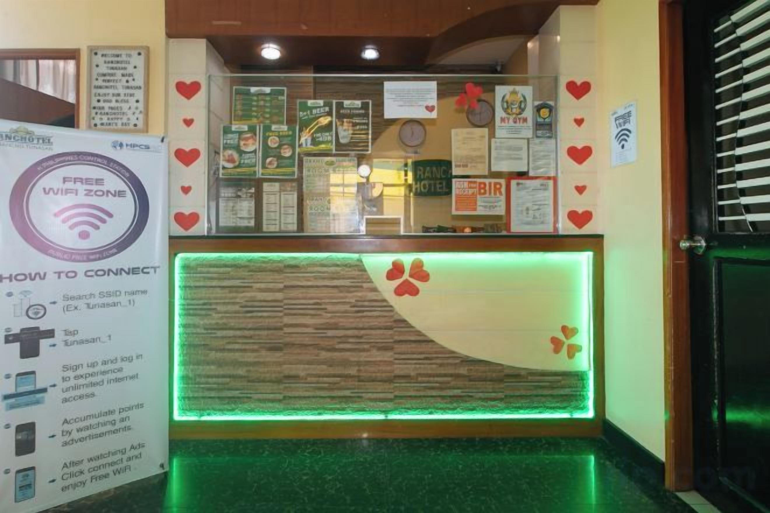 Una reception hotel con un bancone illuminato di verde, cartelli informativi e un segnale che indica 'Free WiFi Zone'.