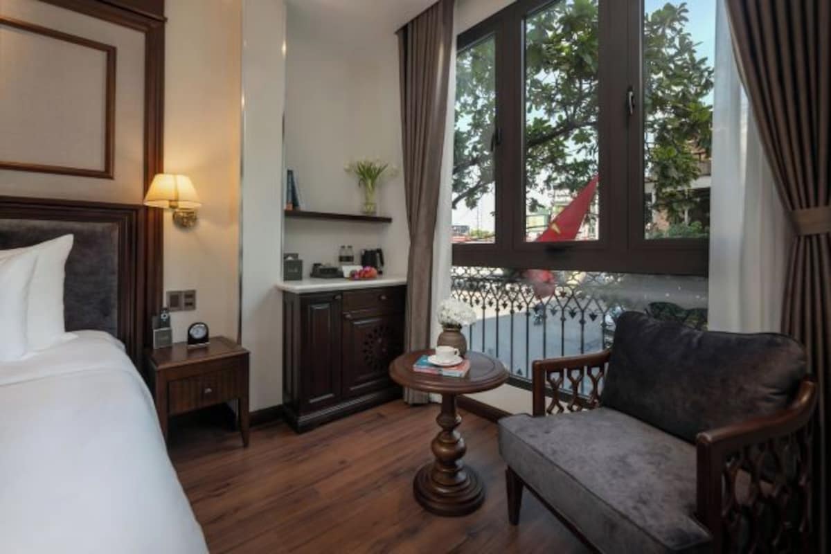A hotel room with a bed, a small table with a cup and book, a chair, a nightstand, a lamp, a window with a view of the outside, and a shelf with decorative items.