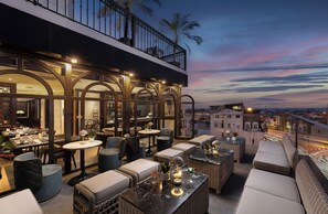 Een buitenontbijtplaats met tafels en stoelen, een balkon met hekwerk en uitzicht op de stad.