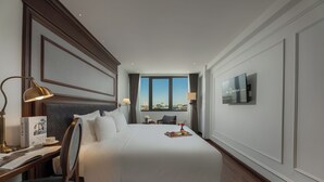 Habitación de hotel con una cama grande, un escritorio con lámpara, televisor en la pared y vistas a la ciudad por la ventana.