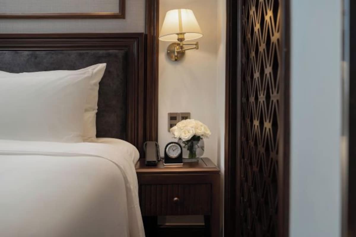 A neatly made bed with white linens, a bedside table with a clock and a vase of white flowers, and a wall-mounted lamp.