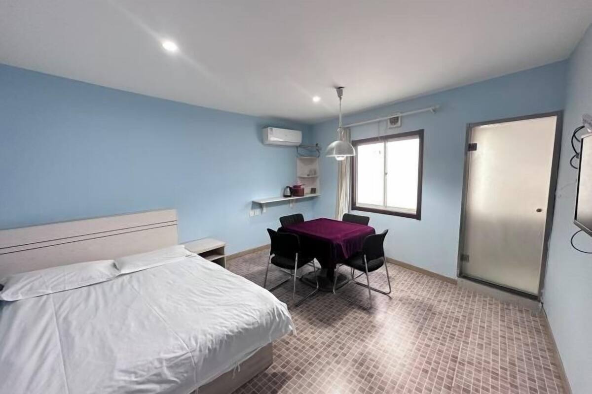 A bedroom with a bed, a table with chairs, a window, and an air conditioning unit.