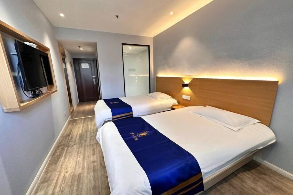 A hotel room with two beds, a TV, a wooden headboard, and a wall-mounted light.