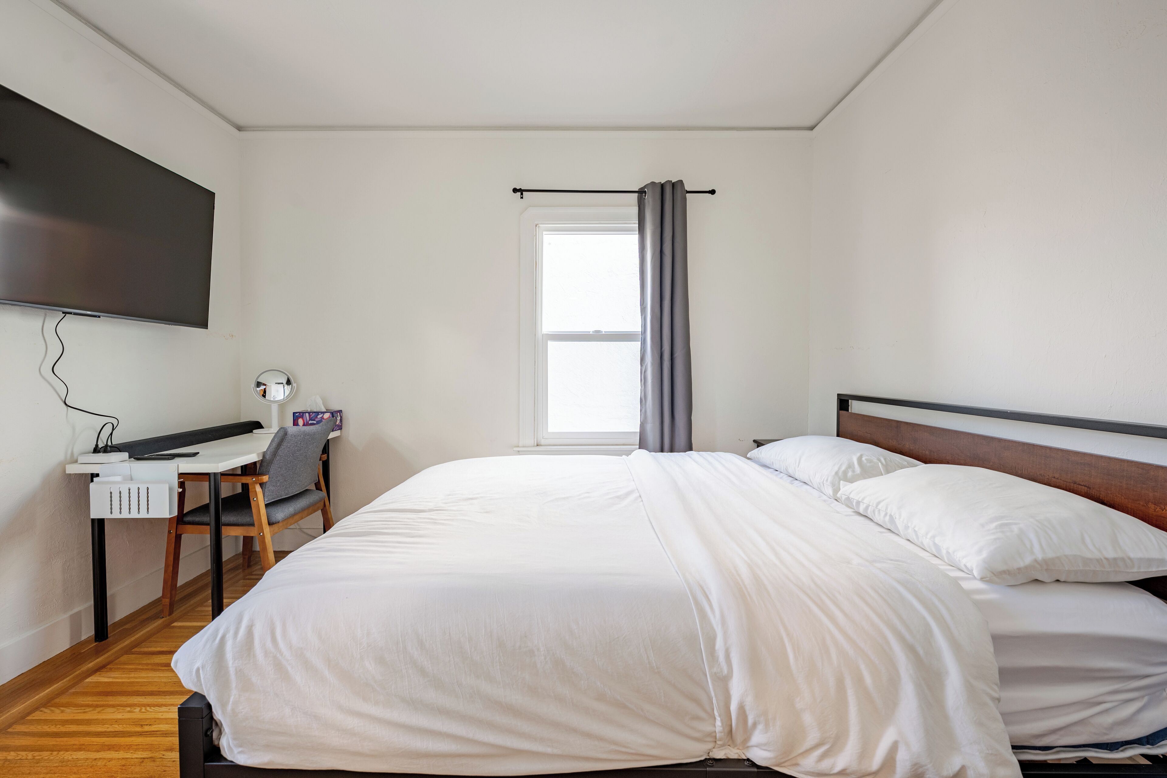 A bedroom with a bed, a desk, a chair, a TV, and a window with curtains.