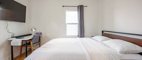 A bedroom with a bed, a desk, a chair, a TV, and a window with curtains.