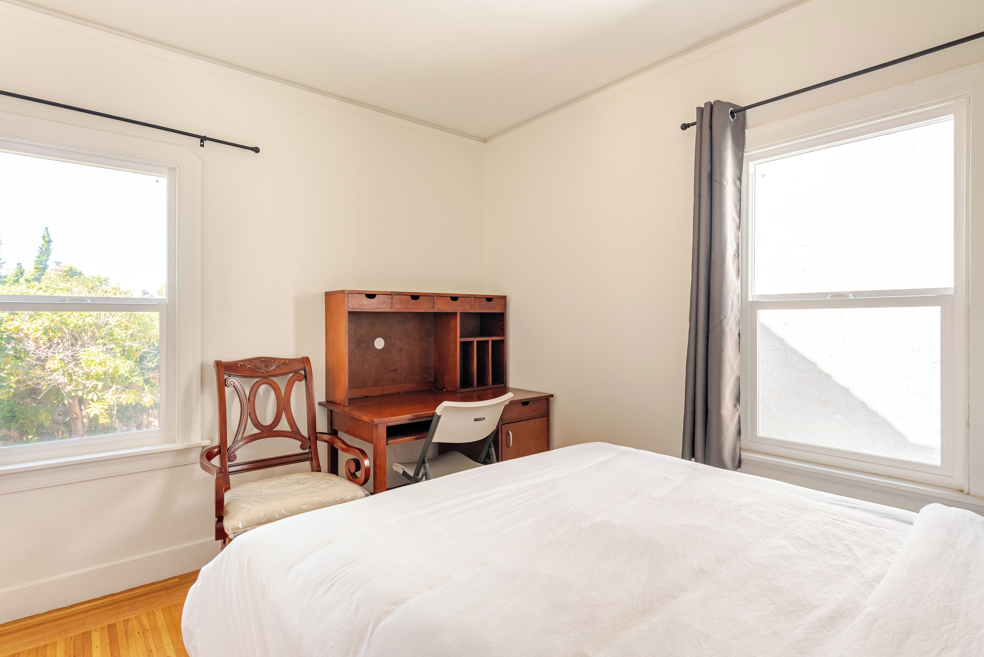 Een slaapkamer met een bed, een houten bureau, een stoel en twee ramen.