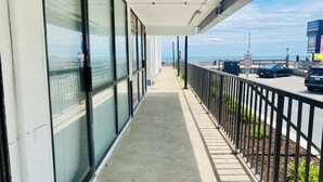 A long, narrow balcony with a view of the sea, a parking area with cars, and a building with large windows.