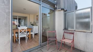Un patio moderno con puerta corrediza de vidrio que da a un área de comedor interior, un pequeño rincón de descanso con dos sillas y un ventanal grande con mosquitero.