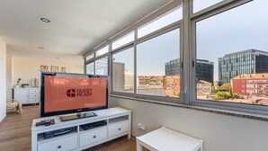 A modern hotel room with a large window, a flat-screen TV displaying "GUEST READY", a white console table, and a small white table.