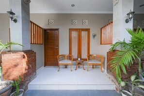 Un espace extérieur abrité, aménagé avec deux chaises en bois et une table, une porte en bois et un pot de fleurs.
